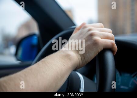 Eine unkenntliche Person hält ihre rechte Hand am Lenkrad und fährt ein modernes Auto mit schwarzem Interieur. Es ist ein Tag in der Stadt draußen. Menschliche Fahrten Stockfoto