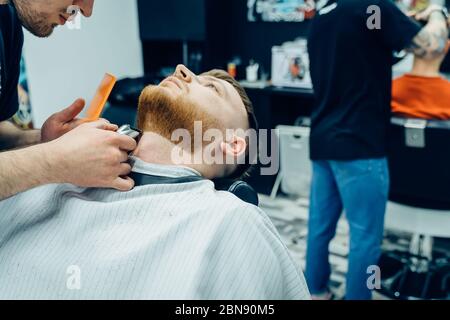 Barber Trimmen bärtiger Mann mit Rasiermaschine im Barbershop. Haarstyling-Prozess. Nahaufnahme eines Haarstylisten, der den Bart eines bärtigen Männchens schneidet. Stockfoto
