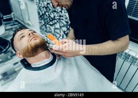 Barber Trimmen bärtiger Mann mit Rasiermaschine im Barbershop. Haarstyling-Prozess. Nahaufnahme eines Haarstylisten, der den Bart eines bärtigen Männchens schneidet. Stockfoto