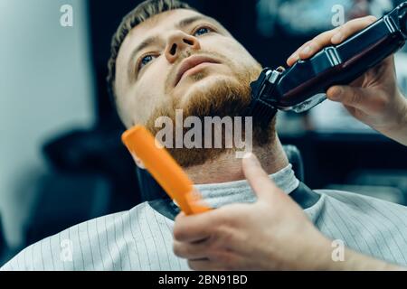 Barber Trimmen bärtiger Mann mit Rasiermaschine im Barbershop. Haarstyling-Prozess. Nahaufnahme eines Haarstylisten, der den Bart eines bärtigen Männchens schneidet. Stockfoto