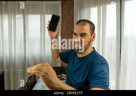 Wütender Mann mit Bart und kurzen Haaren in einem marineblauen Hemd, der sein Handy im Wohnzimmer zu Hause wirft Stockfoto