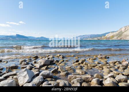 Blick auf die Wellen, die auf den felsigen Strand am Okanagan Lake Rollen, mit Blick auf den blauen Himmel und die Berge Stockfoto