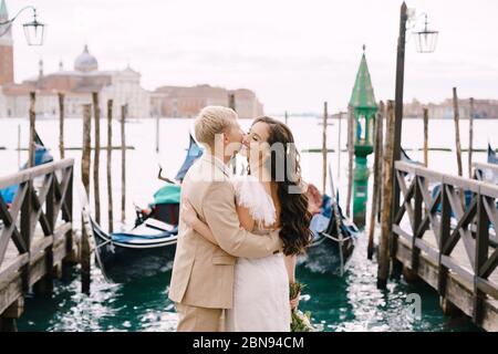 Die Braut und der Bräutigam küssen sich auf der Gondelpier, umarmen sich in Venedig, nahe dem Markusplatz, mit Blick auf San Giorgio Maggiore und den Sonnenuntergang Himmel. Der Stockfoto