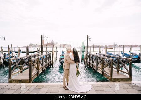 Die Braut und der Bräutigam küssen sich auf der Gondelpier, umarmen sich in Venedig, nahe dem Markusplatz, mit Blick auf San Giorgio Maggiore und den Sonnenuntergang Himmel. Der Stockfoto