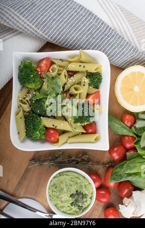 Glutenfreie, frisch gekochte Penne-Pasta aus grünen Erbsen auf Holzbrett isoliert. basilikum, Tomaten, Avocado und Koriander in einer weißen Schüssel. Schuss von oben Stockfoto