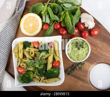 Glutenfreie, frisch gekochte Penne-Pasta aus grünen Erbsen auf Holzbrett isoliert. basilikum, Tomaten, Avocado und Koriander in einer weißen Schüssel. Schuss von oben Stockfoto