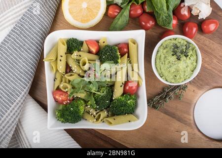 Glutenfreie, frisch gekochte Penne-Pasta aus grünen Erbsen auf Holzbrett isoliert. basilikum, Tomaten, Avocado und Koriander in einer weißen Schüssel. Schuss von oben Stockfoto