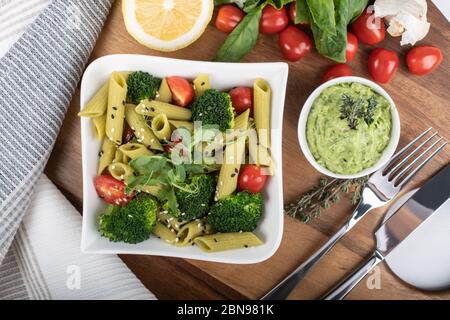Glutenfreie, frisch gekochte Penne-Pasta aus grünen Erbsen auf Holzbrett isoliert. basilikum, Tomaten, Avocado und Koriander in einer weißen Schüssel. Schuss von oben Stockfoto