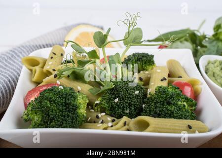 Glutenfreie, frisch gekochte Penne-Pasta aus grünen Erbsen auf Holzbrett isoliert. basilikum, Tomaten, Avocado und Koriander in einer weißen Schüssel. Schuss von oben Stockfoto