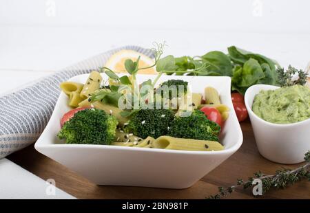 Glutenfreie, frisch gekochte Penne-Pasta aus grünen Erbsen auf Holzbrett isoliert. basilikum, Tomaten, Avocado und Koriander in einer weißen Schüssel. Schuss von oben Stockfoto