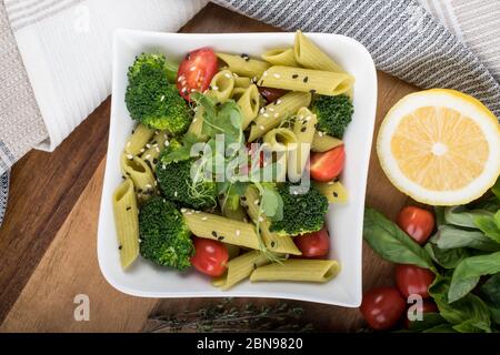 Glutenfreie, frisch gekochte Penne-Pasta aus grünen Erbsen auf Holzbrett isoliert. basilikum, Tomaten, Avocado und Koriander in einer weißen Schüssel. Schuss von oben Stockfoto