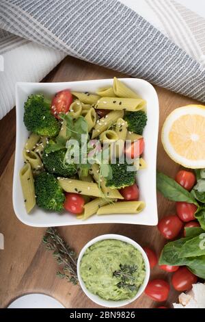 Glutenfreie, frisch gekochte Penne-Pasta aus grünen Erbsen auf Holzbrett isoliert. basilikum, Tomaten, Avocado und Koriander in einer weißen Schüssel. Schuss von oben Stockfoto