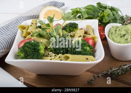 Glutenfreie, frisch gekochte Penne-Pasta aus grünen Erbsen auf Holzbrett isoliert. basilikum, Tomaten, Avocado und Koriander in einer weißen Schüssel. Schuss von oben Stockfoto