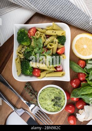 Glutenfreie, frisch gekochte Penne-Pasta aus grünen Erbsen auf Holzbrett isoliert. basilikum, Tomaten, Avocado und Koriander in einer weißen Schüssel. Schuss von oben Stockfoto