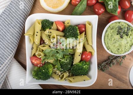 Glutenfreie, frisch gekochte Penne-Pasta aus grünen Erbsen auf Holzbrett isoliert. basilikum, Tomaten, Avocado und Koriander in einer weißen Schüssel. Schuss von oben Stockfoto