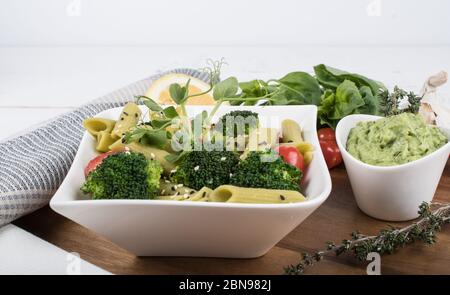 Glutenfreie, frisch gekochte Penne-Pasta aus grünen Erbsen auf Holzbrett isoliert. basilikum, Tomaten, Avocado und Koriander in einer weißen Schüssel. Schuss von oben Stockfoto