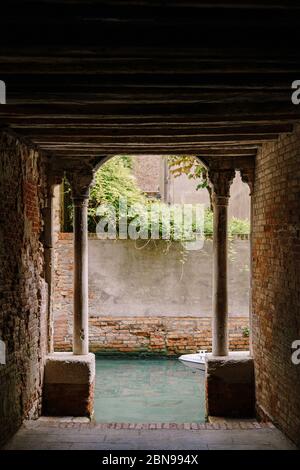 Blick vom Gebäude auf einen kleinen schmalen Kanal zwischen den Häusern in Venedig, Italien. Ein Blick aus einem Tunnel mit Backsteinwänden, zwei antiken Säulen und Stockfoto