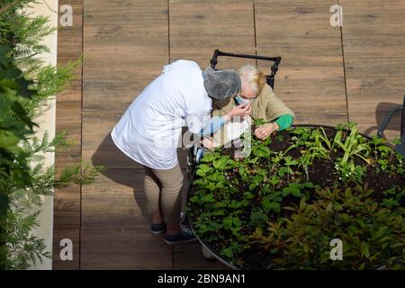 Krankenschwester Pflege einer älteren und behinderten Frau in einem Pflegeheim an einem sonnigen Tag während der Coronavirus-Pandemie Covid-19. Beide tragen eine Gesichtsmaske. Stockfoto