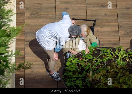 Krankenschwester Pflege einer älteren und behinderten Frau in einem Pflegeheim an einem sonnigen Tag während der Coronavirus-Pandemie Covid-19. Beide tragen eine Gesichtsmaske. Stockfoto