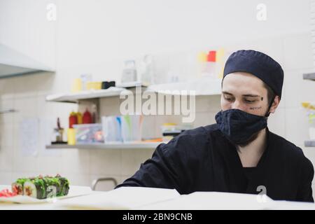 Porträt eines Sushi-Küchenchefs mit Gesichtsmaske in der kommerziellen Küche. Essen zum Mitnehmen, Image des lokalen Catering-Unternehmens Stockfoto