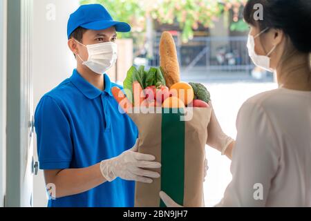 Asiatische Lieferung Mann trägt Gesichtsmaske und Handschuh mit Lebensmitteln Beutel mit Lebensmitteln, Obst, Gemüse geben Frau Kunden vor dem Haus während der Zeit o Stockfoto