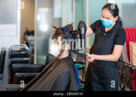 Asiatische junge Frau trägt Gesichtsmasken, um sich von Covid-19 während des Friseurs mit Haartrockner und Kamm zu schwarzen Haaren im Schönheitssalon zu schützen. Stockfoto