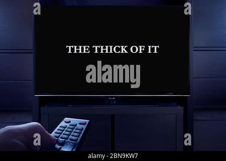 Ein Mann zeigt eine Fernbedienung auf den Fernseher, der den Hauptbildschirm „The Thick of IT“ (nur redaktionelle Verwendung) anzeigt. Stockfoto