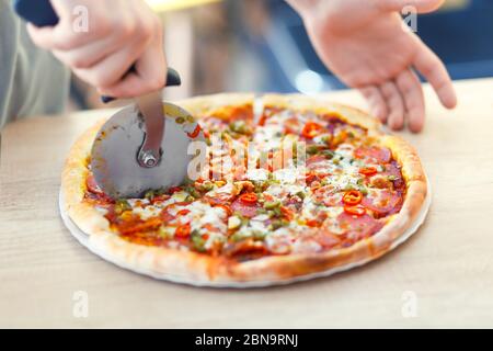 Chefbäcker in einheitlicher Form, der frische Pizzaböcker auf Holzbrett in der Küche schneidet, Soft Focus. Hand schneiden eine Pepperoni Pizza mit Cutter auf dem Tisch. Stockfoto