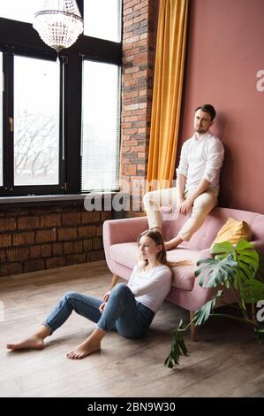 Mann sitzt auf dem Rücken des Sofas und schaut auf die Kamera, Frau sitzt auf dem Boden in der Nähe. Stockfoto