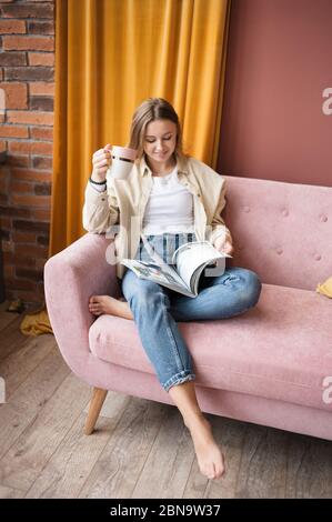Frau sitzt auf rosa Sofa im Wohnzimmer mit Magazin und Tasse Tee. Stockfoto