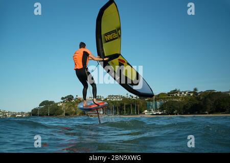 Ein junger Mann wingfoils in Auckland Hafen an einem Sommertag, mit einem Hand gehalten aufblasbaren Flügel und Reiten ein Tragflächenboot Surfboard. Stockfoto