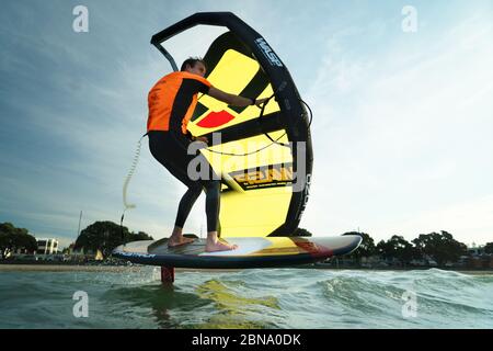 Ein junger Mann wingfoils in Auckland Hafen an einem Sommertag, mit einem Hand gehalten aufblasbaren Flügel und Reiten ein Tragflächenboot Surfboard. Stockfoto