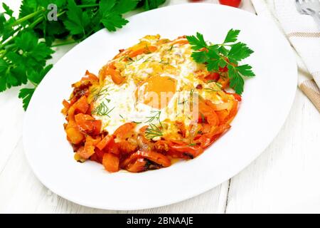 Spiegeleier mit Tomaten, Paprika, Zwiebeln und Kräutern in einem Teller, Serviette, Petersilie und Gabel auf Holzbrett Hintergrund Stockfoto