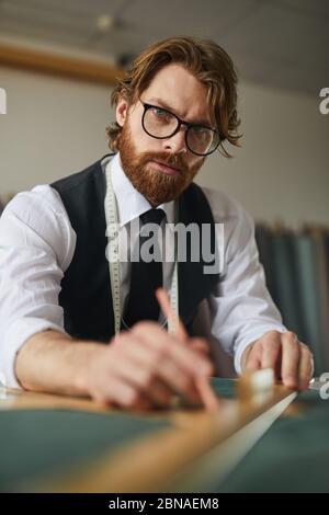 Porträt von bärtigen Schneider in Brillen Blick auf die Kamera, während die Arbeit mit Stoff am Tisch Stockfoto