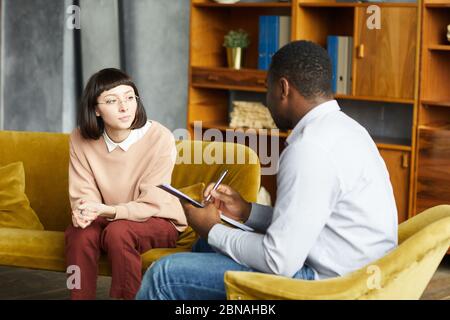 Junge Frau auf dem Sofa sitzen und mit afrikanischen Mann sprechen, während er Notizen in Dokument im Büro Stockfoto