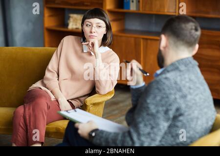 Junge Frau in Brillen, die auf dem Sofa sitzt und mit dem Mann während ihres Besuchs in der Psychotherapie spricht Stockfoto