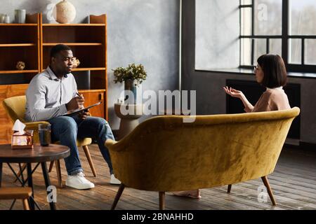 Afrikanische Psychologin sitzt auf Stuhl und spricht mit jungen Frau Sie diskutieren einige Probleme während der Psychotherapie Stockfoto