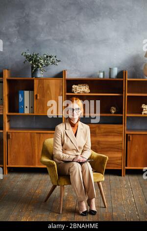 Porträt von rothaarigen reifen Geschäftsfrau in Brillen sitzen auf Sessel und lächeln an Kamera im Büro Stockfoto