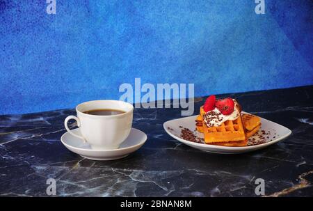 Eine Tasse schwarzen Kaffee auf einer Untertasse und ein Teller mit zwei Wiener Waffeln mit Erdbeeren, Sahne und Schokoladenstreuseln. Nahaufnahme. Stockfoto