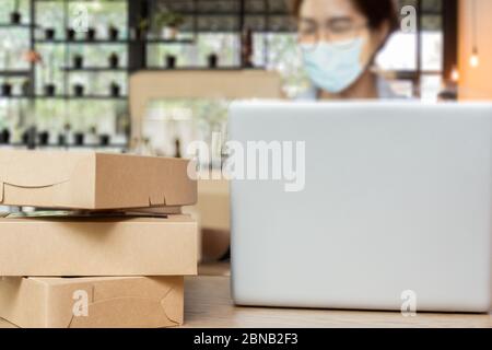 Ausgewählte Fokus Paketbox auf Tisch mit Frau trägt medizinische Maske Arbeit auf Laptop zu Hause. Stockfoto