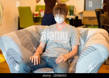 Junger Mann mit Maske mit langen Haaren aufgrund von Corona-Virus-Pandemie, während in Quarantäne zu Hause Stockfoto