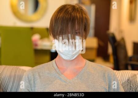 Gesicht des jungen Mannes mit Maske mit langen Haaren aufgrund von Corona-Virus-Pandemie, während in Quarantäne zu Hause Stockfoto