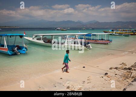 Horizontale Ansicht eines indonesischen Jungen, der am Strand entlang läuft, in der Nähe einiger traditioneller Fischerboote, die in Touristenboote umgewandelt wurden, Gili Meno Stockfoto