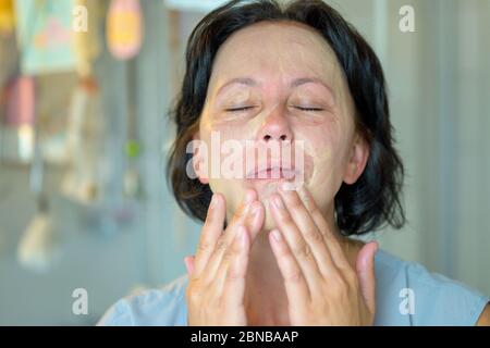 Hispanische Frau, die eine Gesichtsmaske auf ihre Haut aufbringt und sie mit geschlossenen Augen und einem ruhigen Ausdruck in der Nähe in einer Hautpflege einmassiert Stockfoto