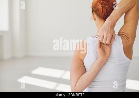 Rückansicht der jungen fit Frau in weißer Sportbekleidung sitzt in Gomukhasana Pose mit den Händen hinter dem Rücken während Yoga in Licht Studio üben Stockfoto