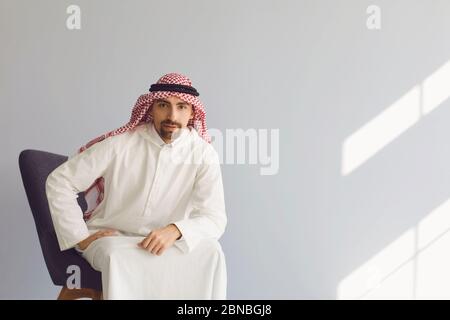 Nachdenklicher ernst arabischer Geschäftsmann, der auf einem Stuhl sitzt, denkt, er sieht auf einem grauen Hintergrund auf. Porträt eines attraktiven arabischen Mannes. Stockfoto