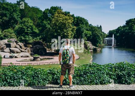 Sofia Park, Ukraine. Mann mit Rucksack in einem Landschaftsgarten im Sommer. Der Mensch drehte den Rücken auf dem Hintergrund des Sees mit einem Brunnen. Stockfoto