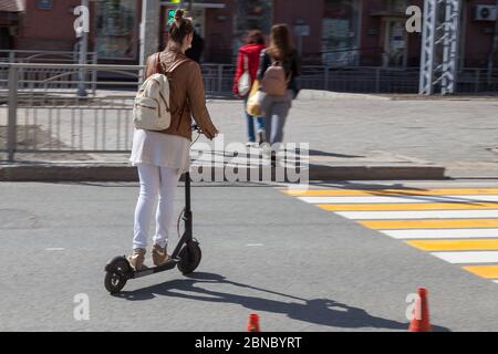 Ein blondes Mädchen in einer Lederjacke und beiger Hose auf einem Elektroroller mit einem Rucksack auf dem Rücken überquert die Straße bei einer Fußgängerüberfahrt Stockfoto