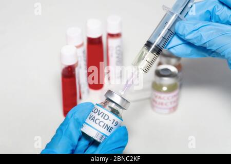 Hände eines Arztes in medizinischen Handschuhen, die eine hypodermische Nadel halten, die Medikamente aus einer Impfstoffflasche mit Blick auf Arzneimittel und Blutproben injiziert Stockfoto