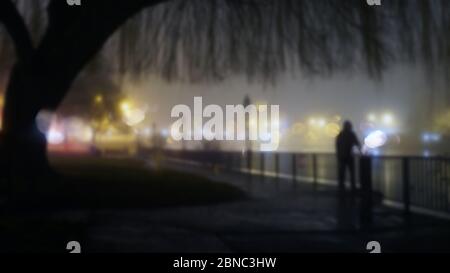 Eine geheimnisvolle Kapuzenfigur, die in einer nebligen Winternacht an Straßenlaternen neben einem Fluss steht. Mit einem unscharfen, Bokeh, unscharf bearbeitenden Schnitt Stockfoto